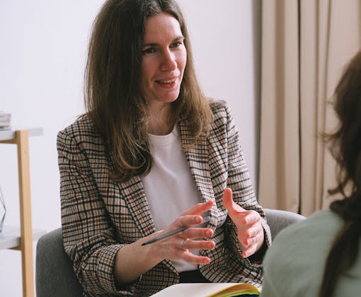 Pensive woman psychologist with brown hair in stylish clothes sitting and talking with unrecognizable female in light room