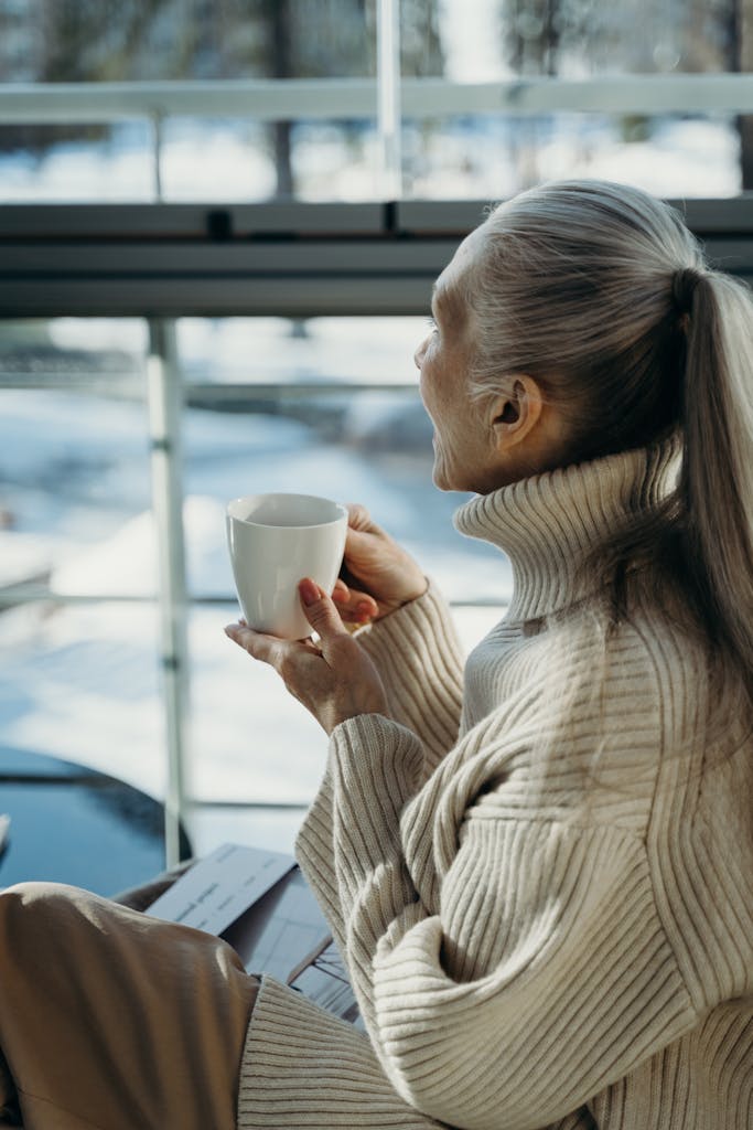 Senior woman in a cozy sweater holding a cup, relaxing by a window on a winter day.
