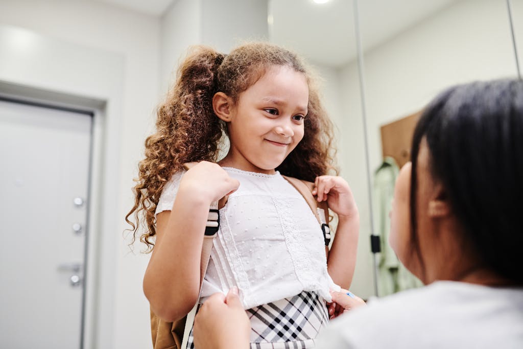 A smiling young girl with curly hair ready for school with her mother indoors.