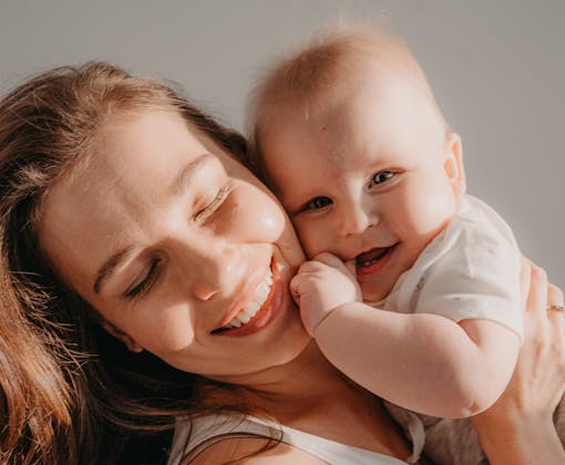 A joyful mother lovingly hugs her smiling baby, both glowing in warm sunlight.