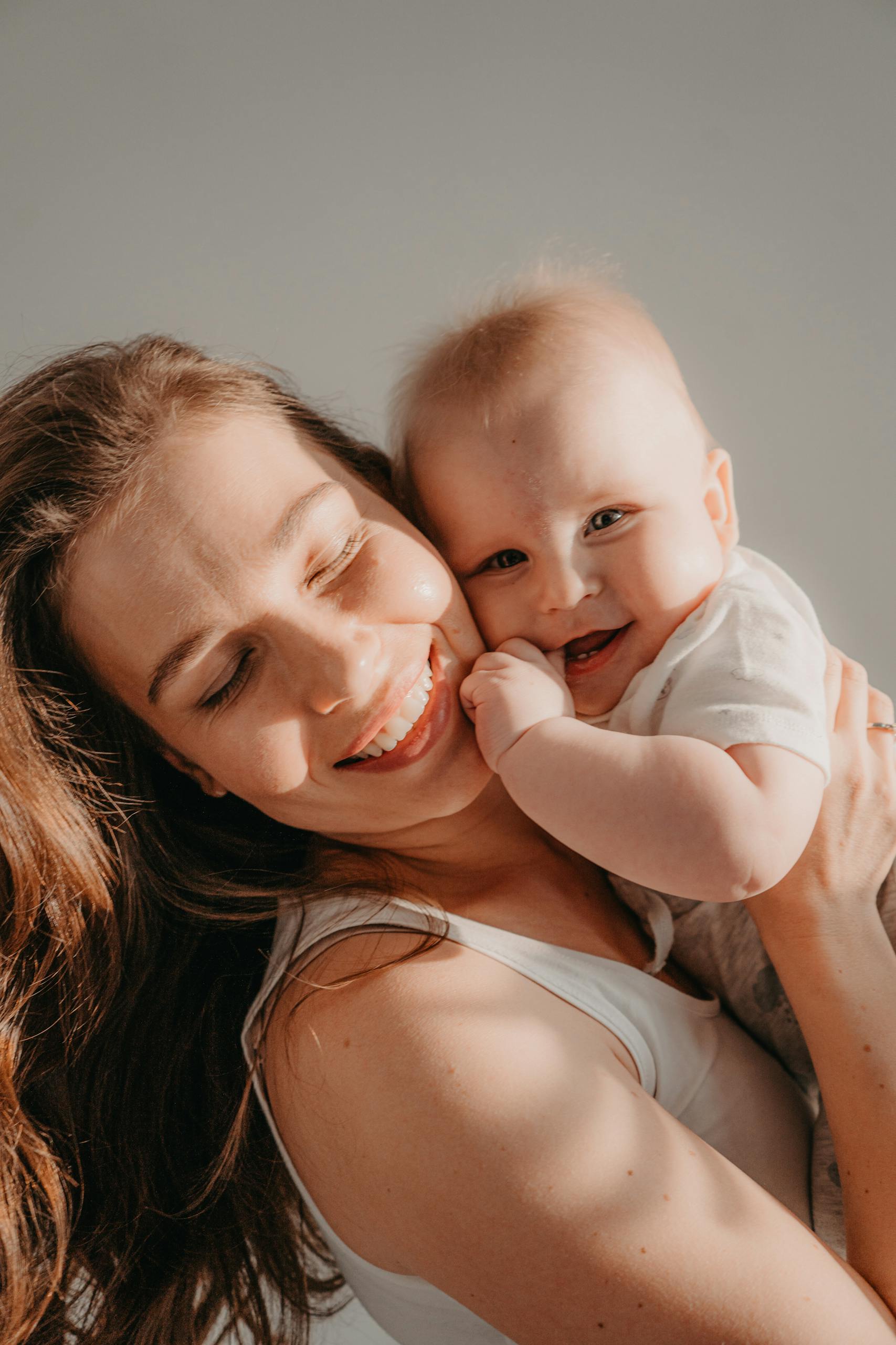 A joyful mother lovingly hugs her smiling baby, both glowing in warm sunlight.