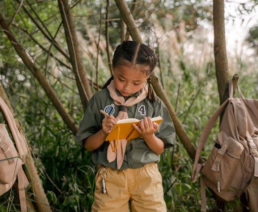 A young girl scout takes notes during a summer camp in the woods.