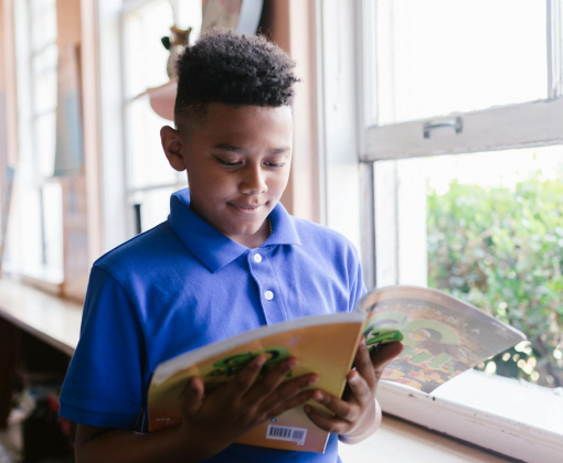 A boy in a blue polo reads a book by a sunlit window, enjoying a quiet moment indoors.