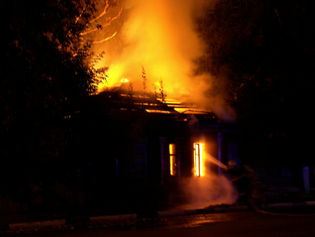 Firefighters battle a raging house fire at night, with smoke and flames spiraling upward.