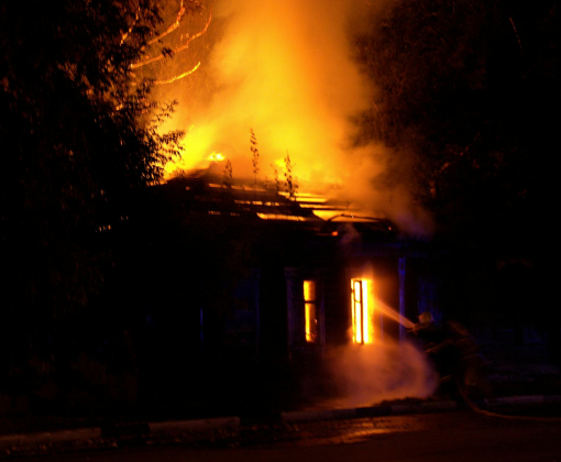 Firefighters battle a raging house fire at night, with smoke and flames spiraling upward.