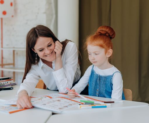 A smiling teacher assists a young girl with a colorful educational activity at a classroom table.