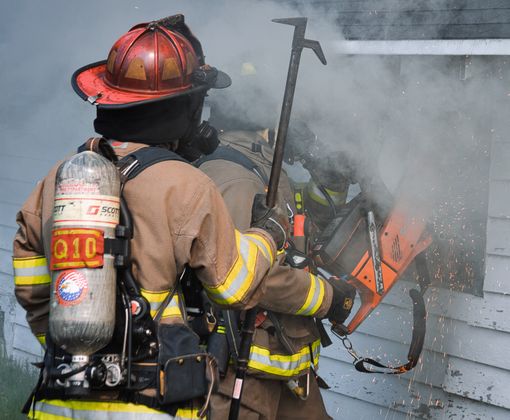 Firefighters using a power tool to cut through smoke-filled building exterior.
